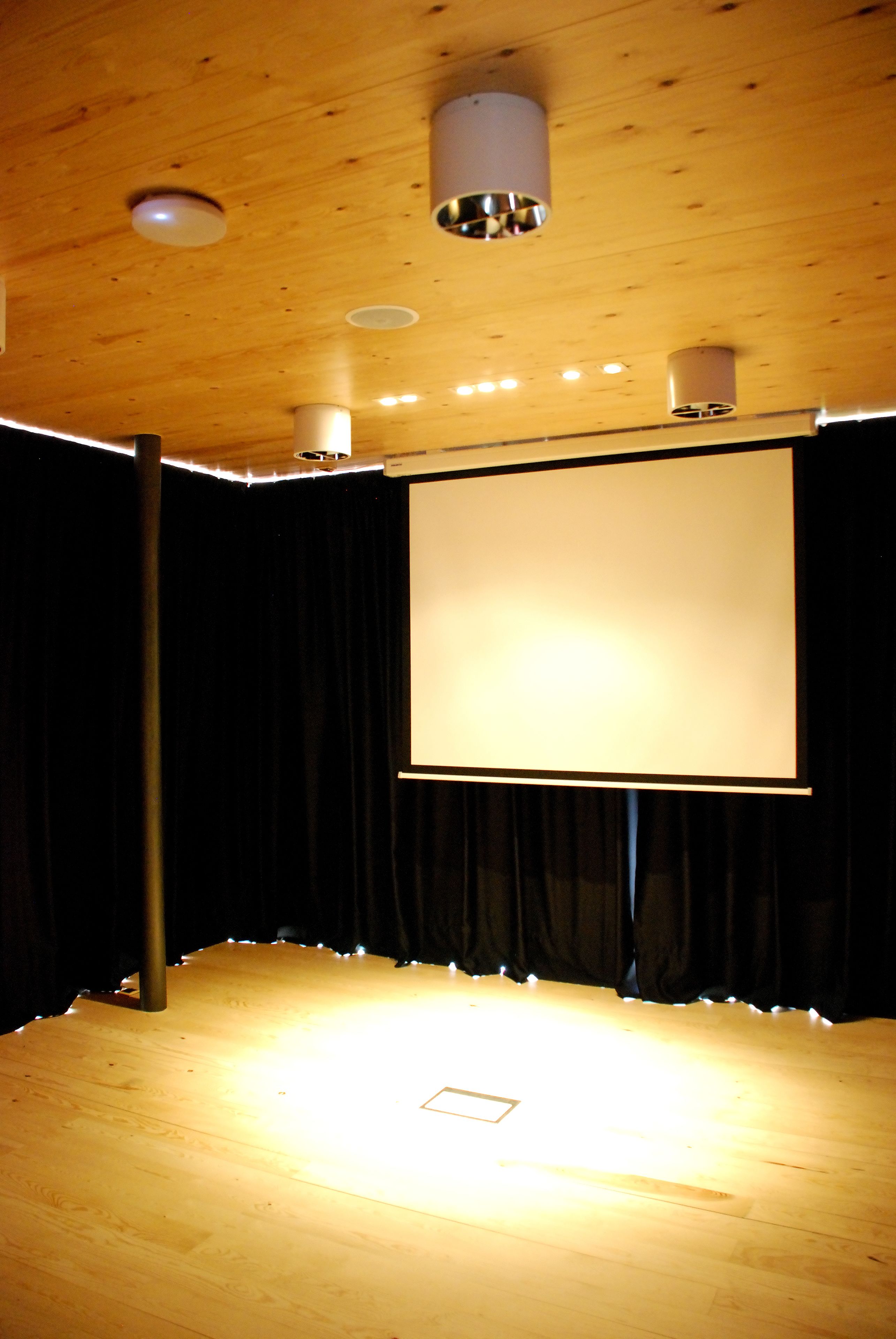 Un teatro con techo de madera, una pantalla grande y cortinas negras.
