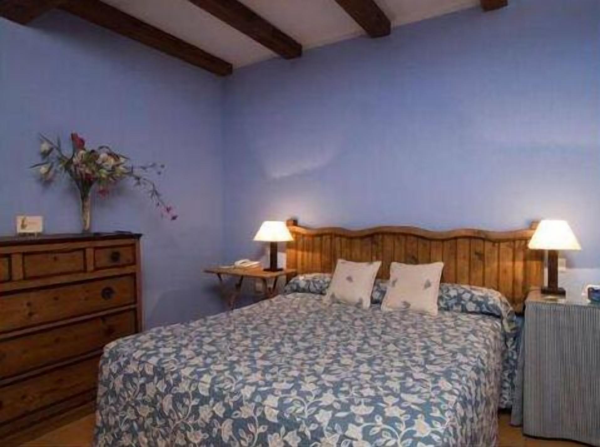 Ein Schlafzimmer mit einem Holzkopfteil, einer blumigen Bettdecke, zwei Nachttischlampen und einer Kommode mit einem Blumenstrauß.