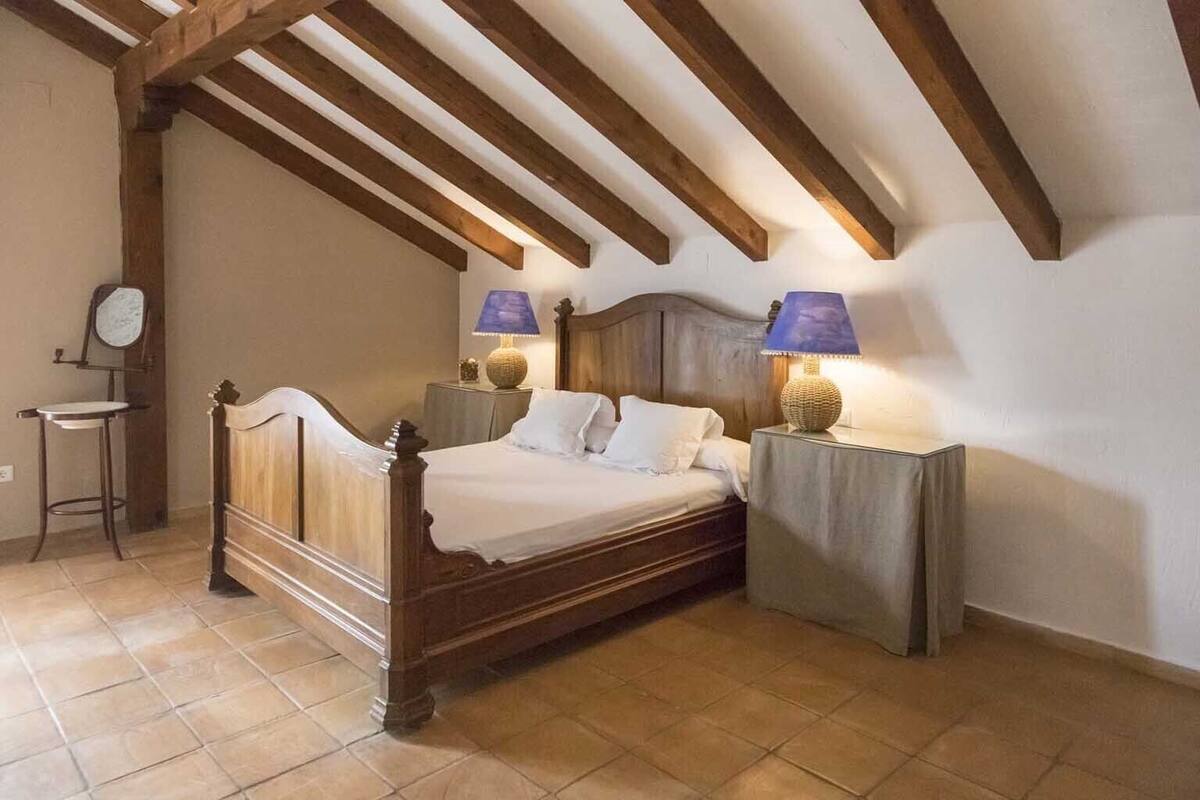 Un dormitori amb un llit de fusta, dues taules de nit amb làmpades, un tamboret petit i un mirall a la paret.