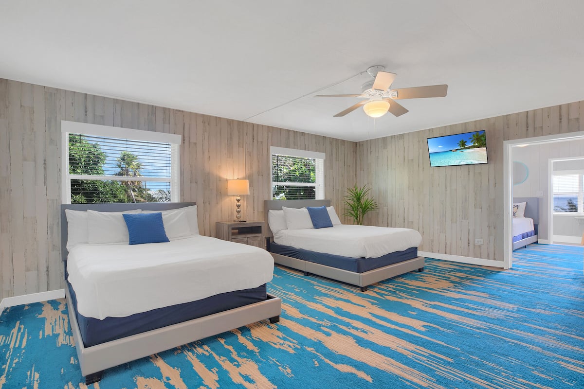 A hotel room with two beds, a ceiling fan, a TV, and a view of the outdoors through the windows.
