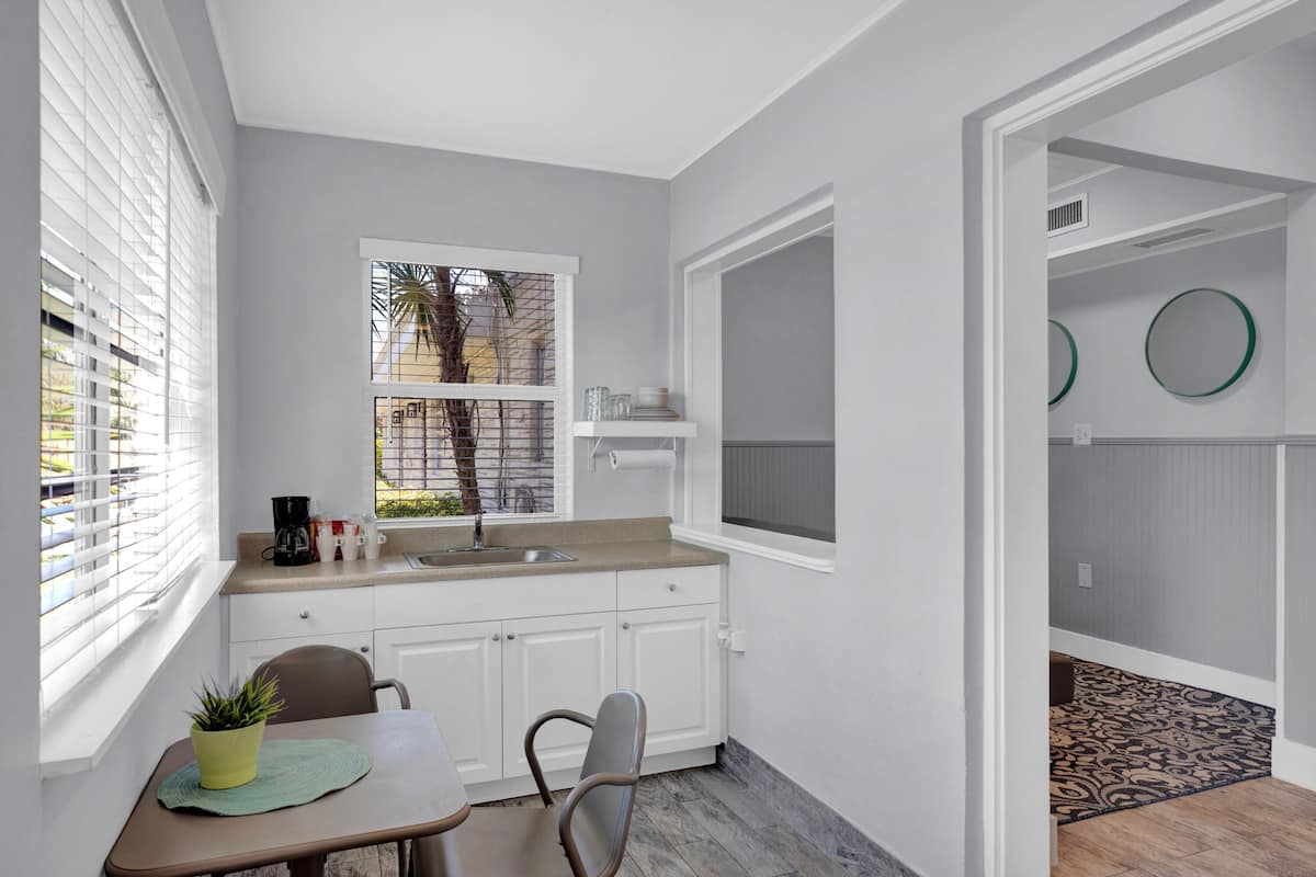 A compact kitchen with white cabinets, a sink, and a window with blinds. There is a small dining table with chairs and a potted plant.