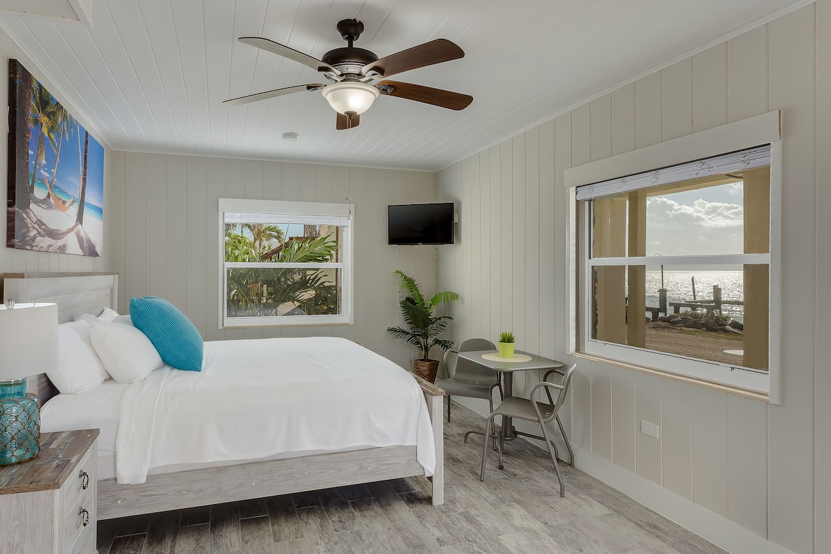 A bedroom with a bed, a ceiling fan, a TV, a window with a view of the beach, and a small table with chairs.