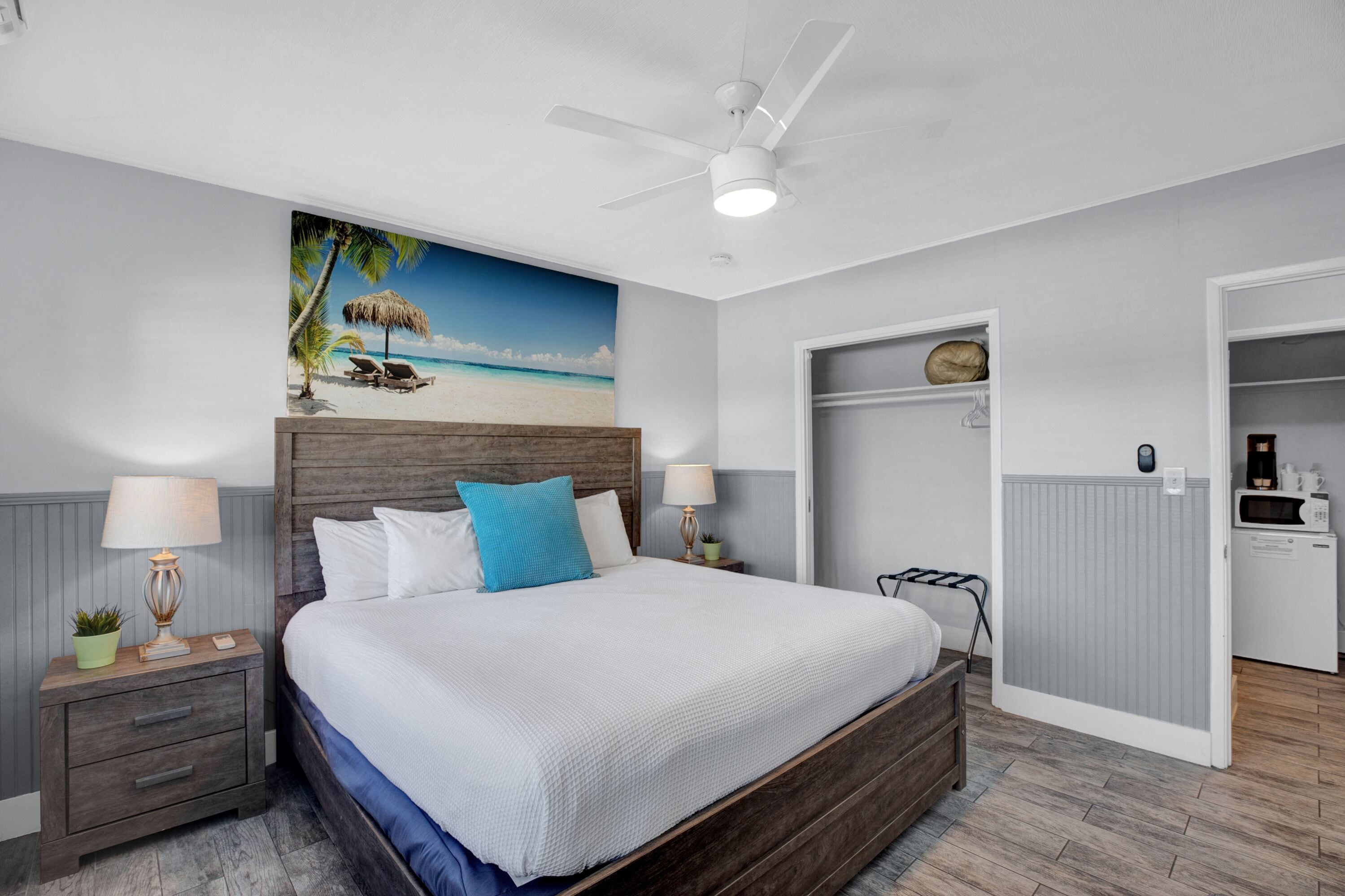 Un dormitorio con cama, mesitas de noche, ventilador de techo, un mural temático de playa, armario y kitchenette.