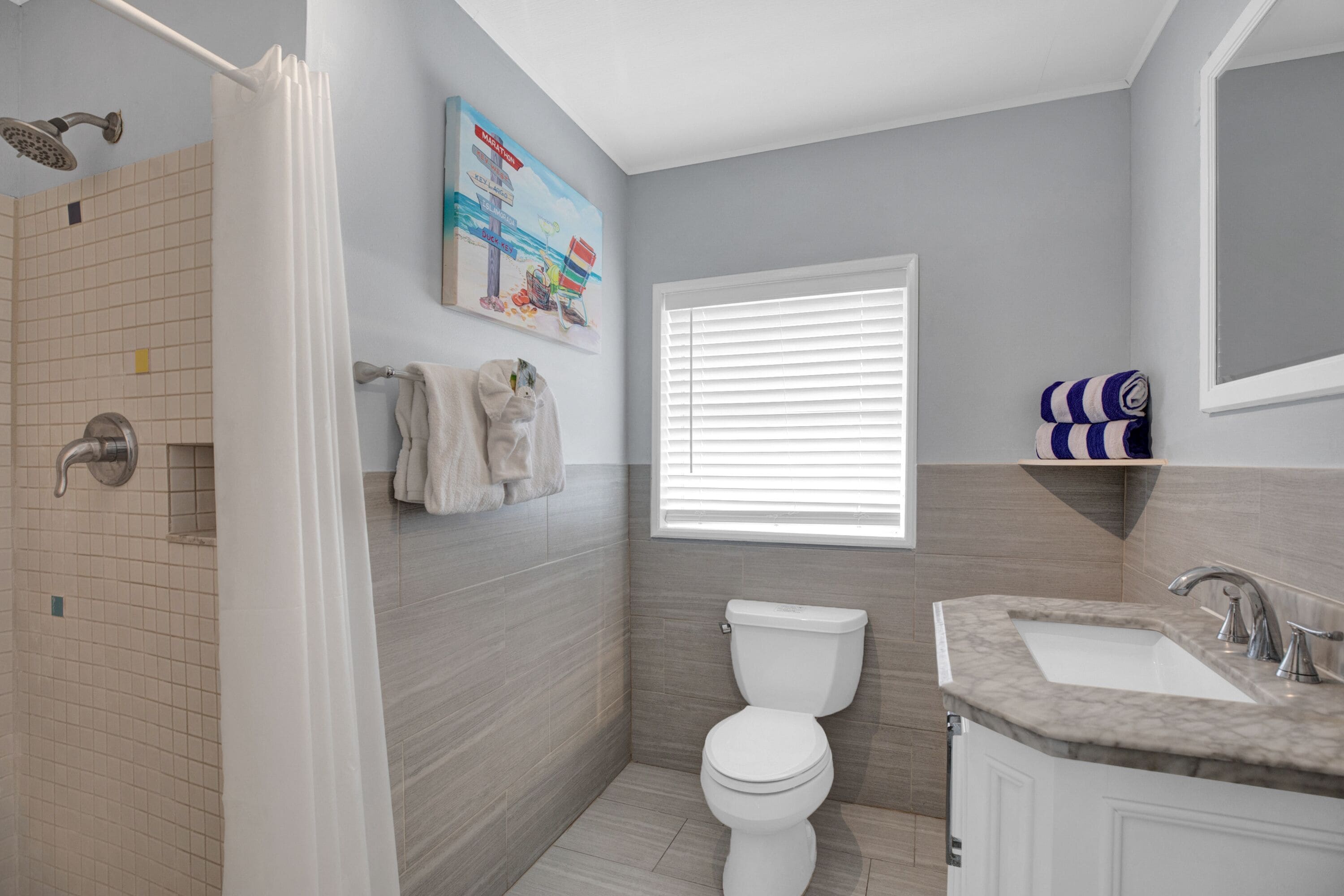A bathroom with a shower, toilet, sink, and a window with blinds.