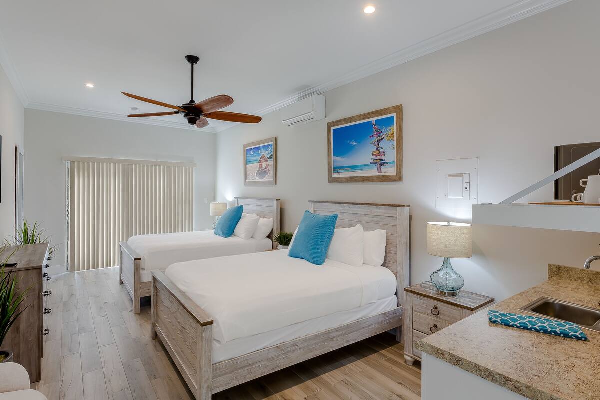 A hotel room with two beds, a ceiling fan, a TV, a desk with a lamp, and artwork on the walls.