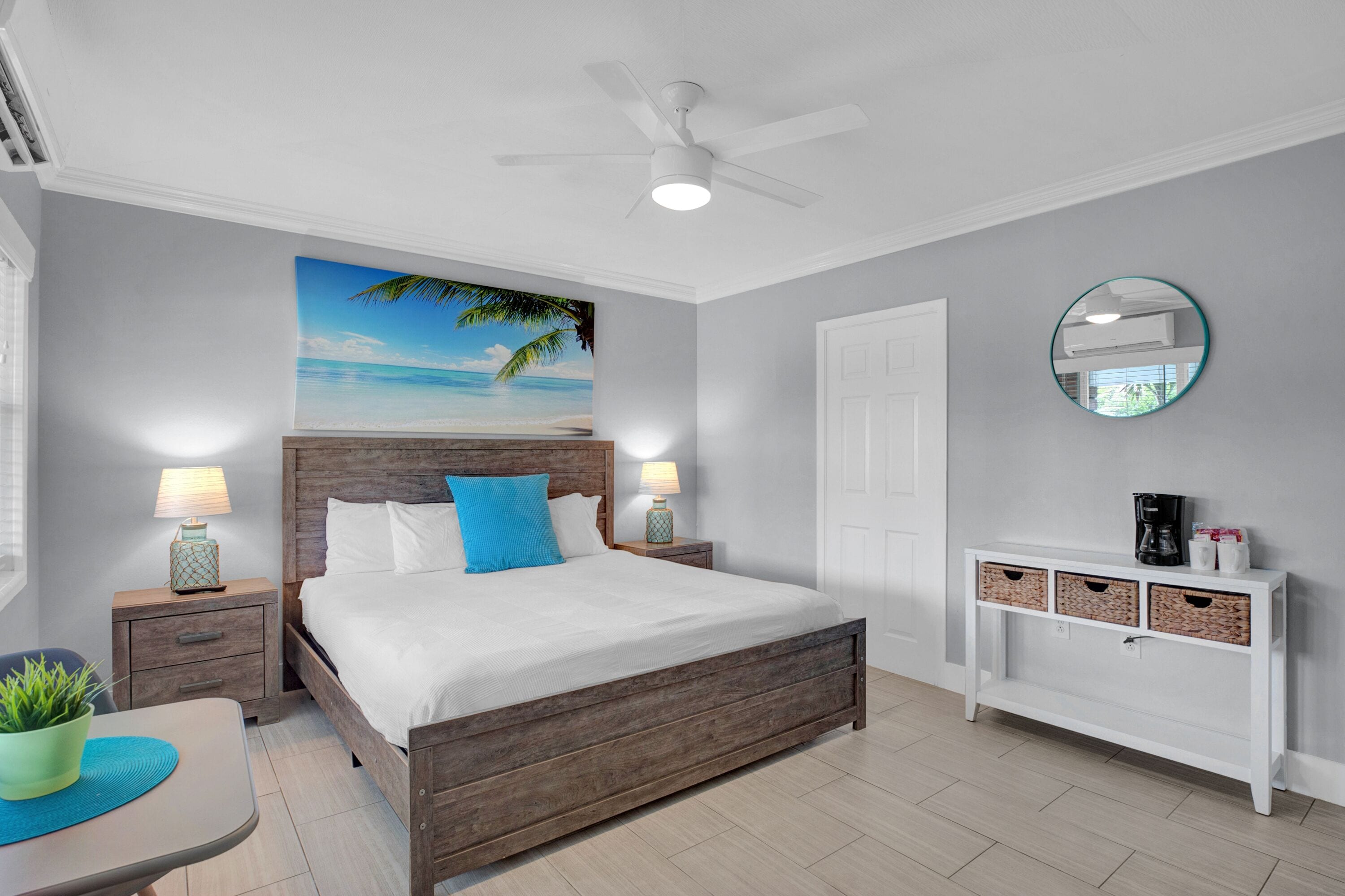 Quarto com cama de madeira, duas mesas de cabeceira com luminárias, um console branco com cestos, um espelho redondo e um ventilador de teto.