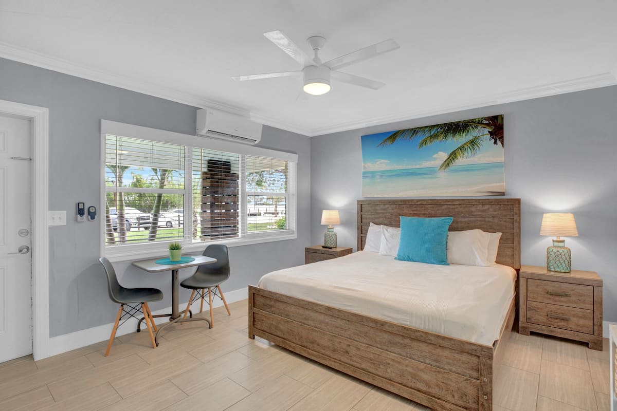 A bedroom with a wooden bed, a desk, two chairs, a ceiling fan, and a beach-themed painting.