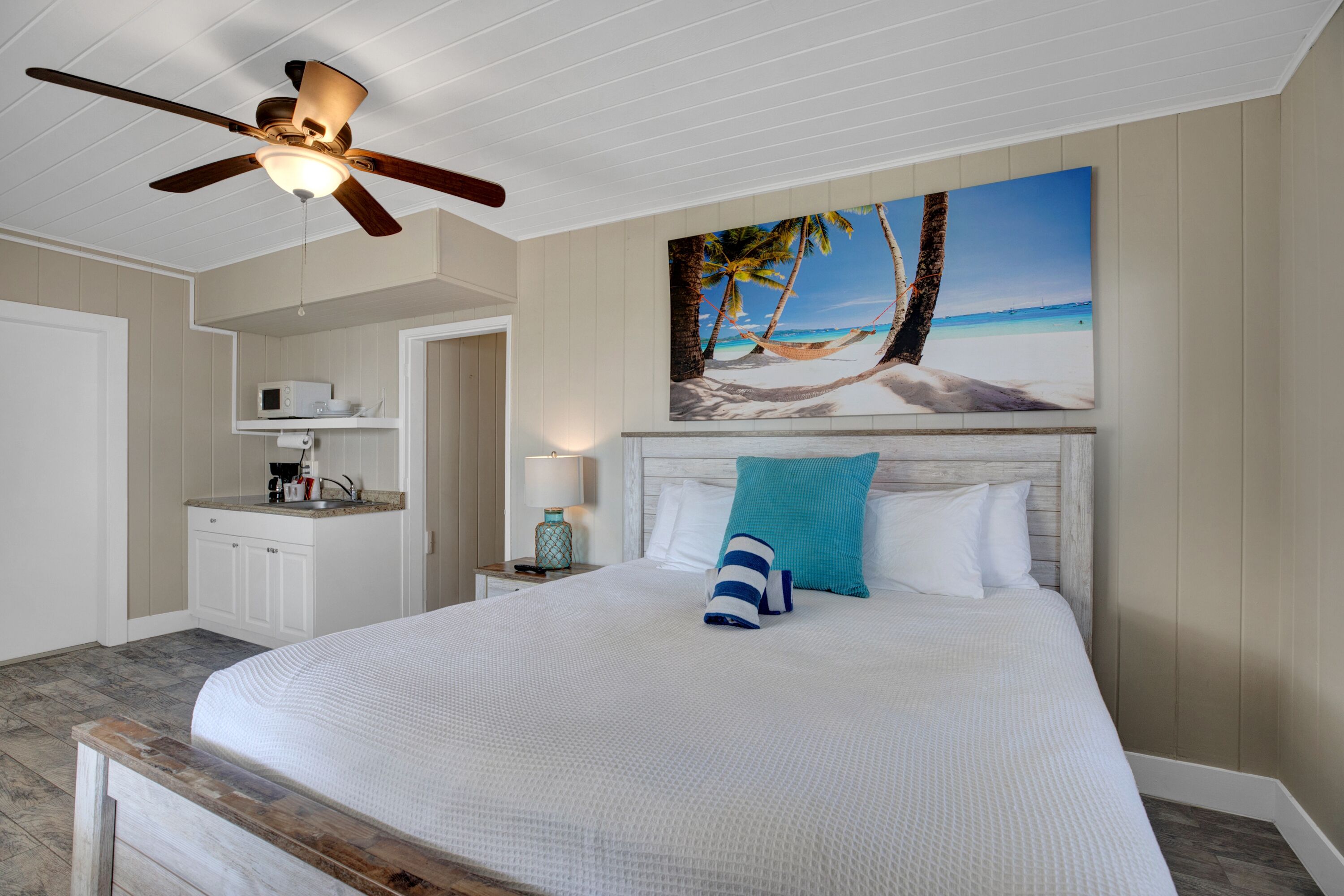 Une chambre à coucher comprenant un lit, un ventilateur de plafond, un tableau représentant une scène de plage, un four à micro-ondes et une table de chevet avec une lampe.