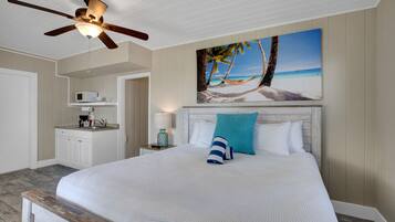 Une chambre à coucher comprenant un lit, un ventilateur de plafond, un tableau représentant une scène de plage, un four à micro-ondes et une table de chevet avec une lampe.