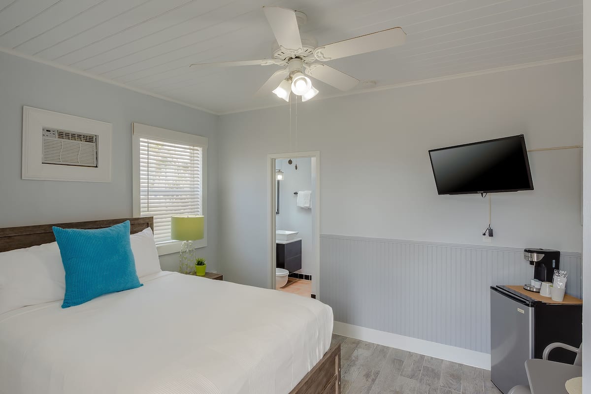 A bedroom with a bed, a ceiling fan, a TV, a radiator, and a small kitchenette.