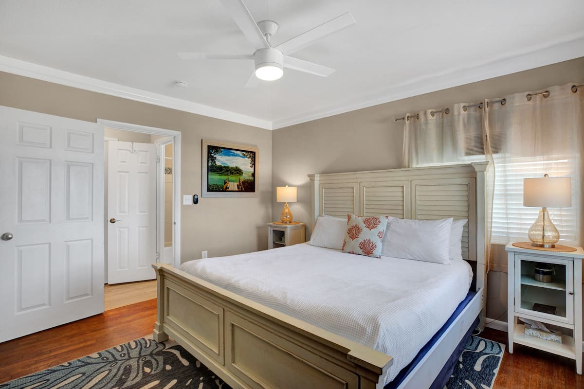 A bedroom with a bed, a ceiling fan, a nightstand with a lamp, a door, and a painting on the wall.