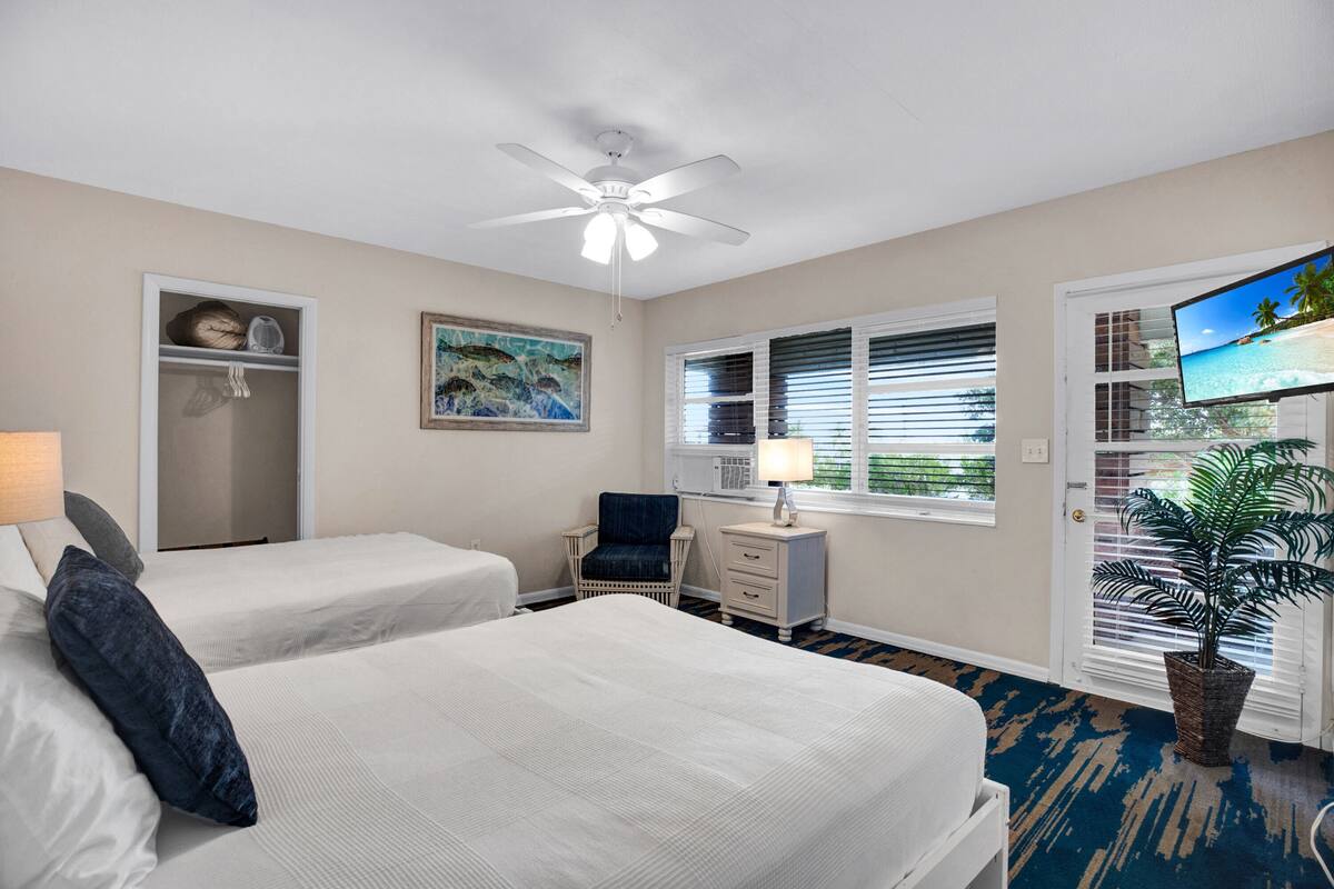 A hotel room with two beds, a ceiling fan, a TV, a window with blinds, a desk with a lamp, a chair, a plant, and a door with a view of the beach.