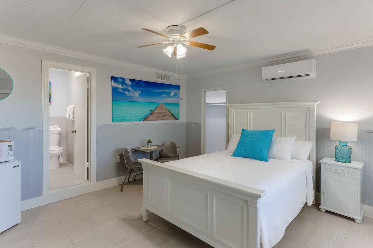 A bedroom with a bed, a ceiling fan, a painting of a beach, a bathroom, and a desk with a chair.