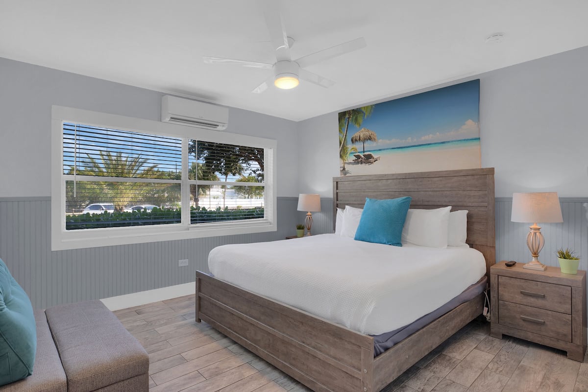 A bedroom with a large bed, a ceiling fan, a window with blinds, a nightstand with a lamp, and a painting of a beach scene.