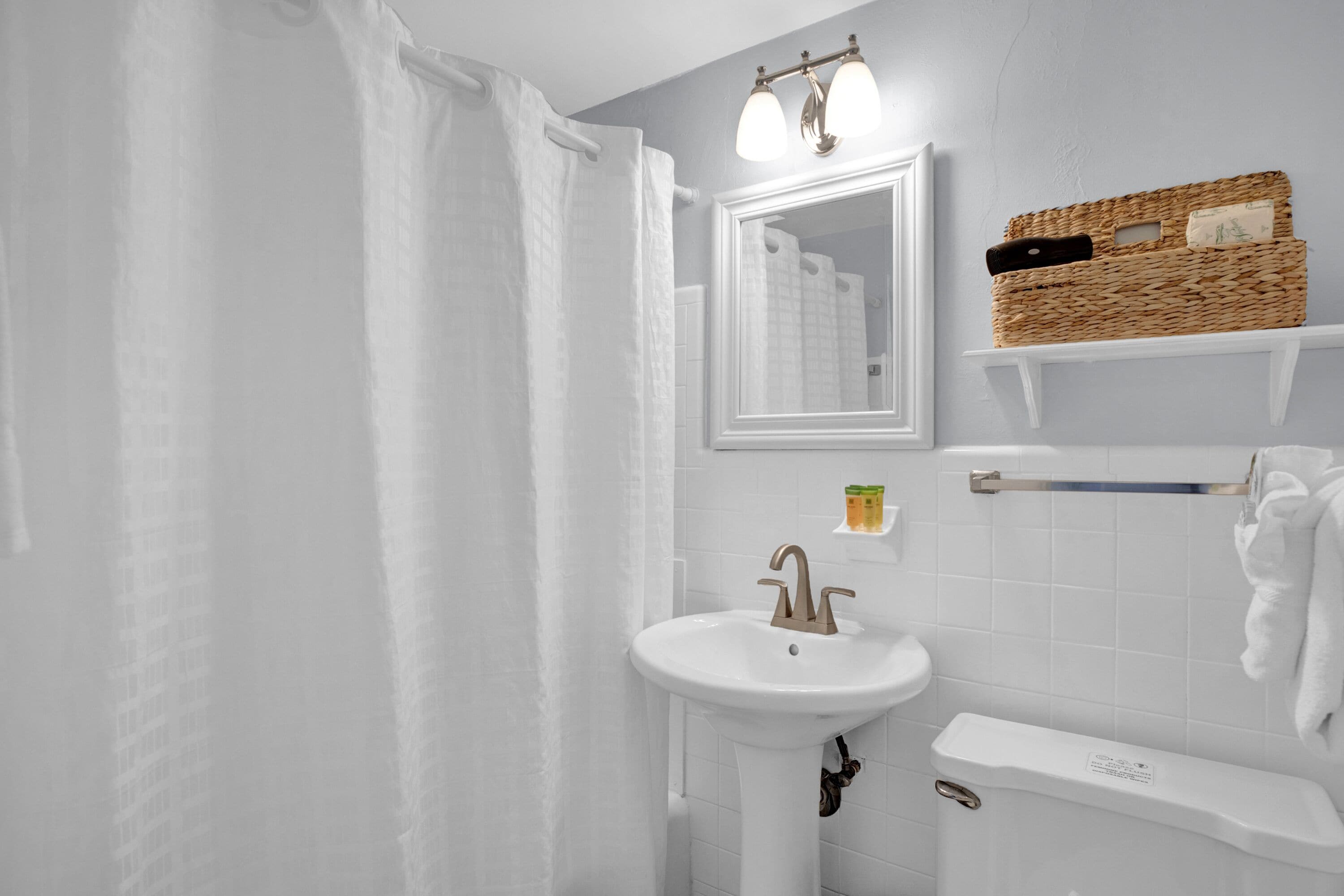 A bathroom with a white sink, a mirror, a shower curtain, a towel rack, and a wicker basket on a shelf.
