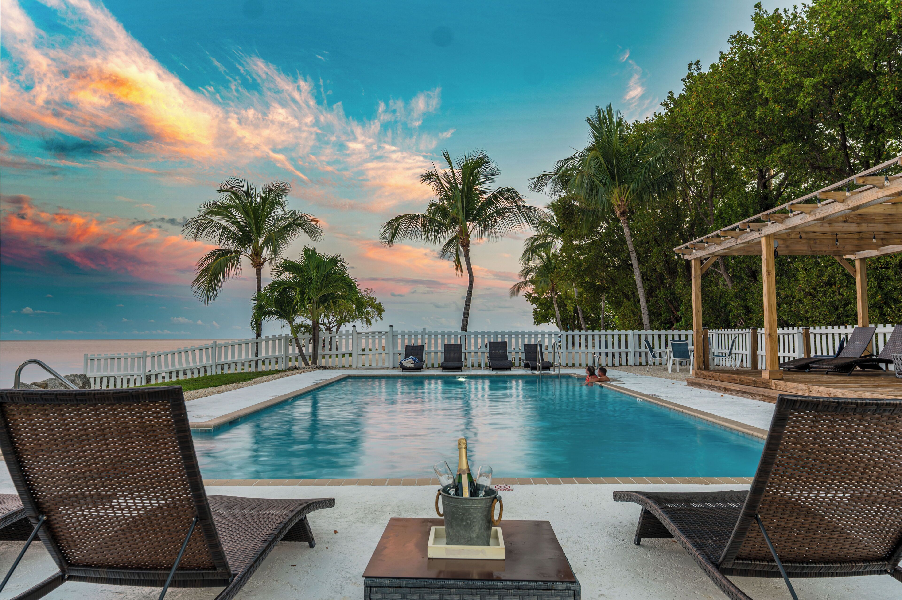 A swimming pool with lounge chairs, a wooden pergola, and a view of the ocean.