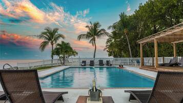 A swimming pool with lounge chairs, a wooden pergola, and a view of the ocean.