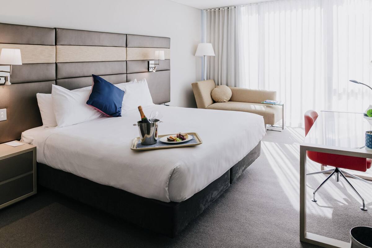 A hotel room with a large bed, a bedside table, a nightstand, a desk with a chair, a lamp, and a window with curtains.