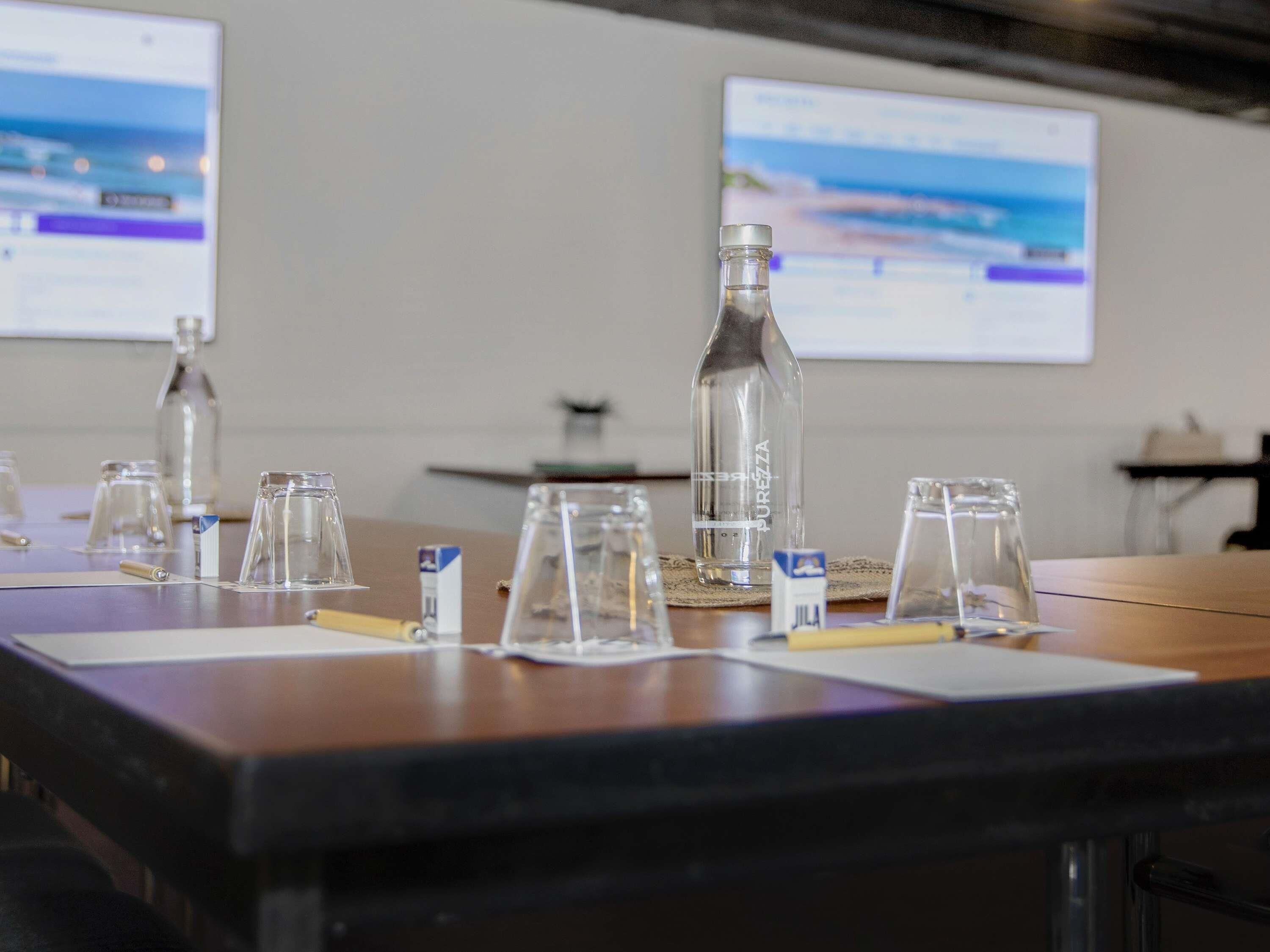 Een vergaderruimte met een tafel, glazen, een fles en een doos met de tekst 'JILA', met twee schermen waarop informatie wordt getoond.