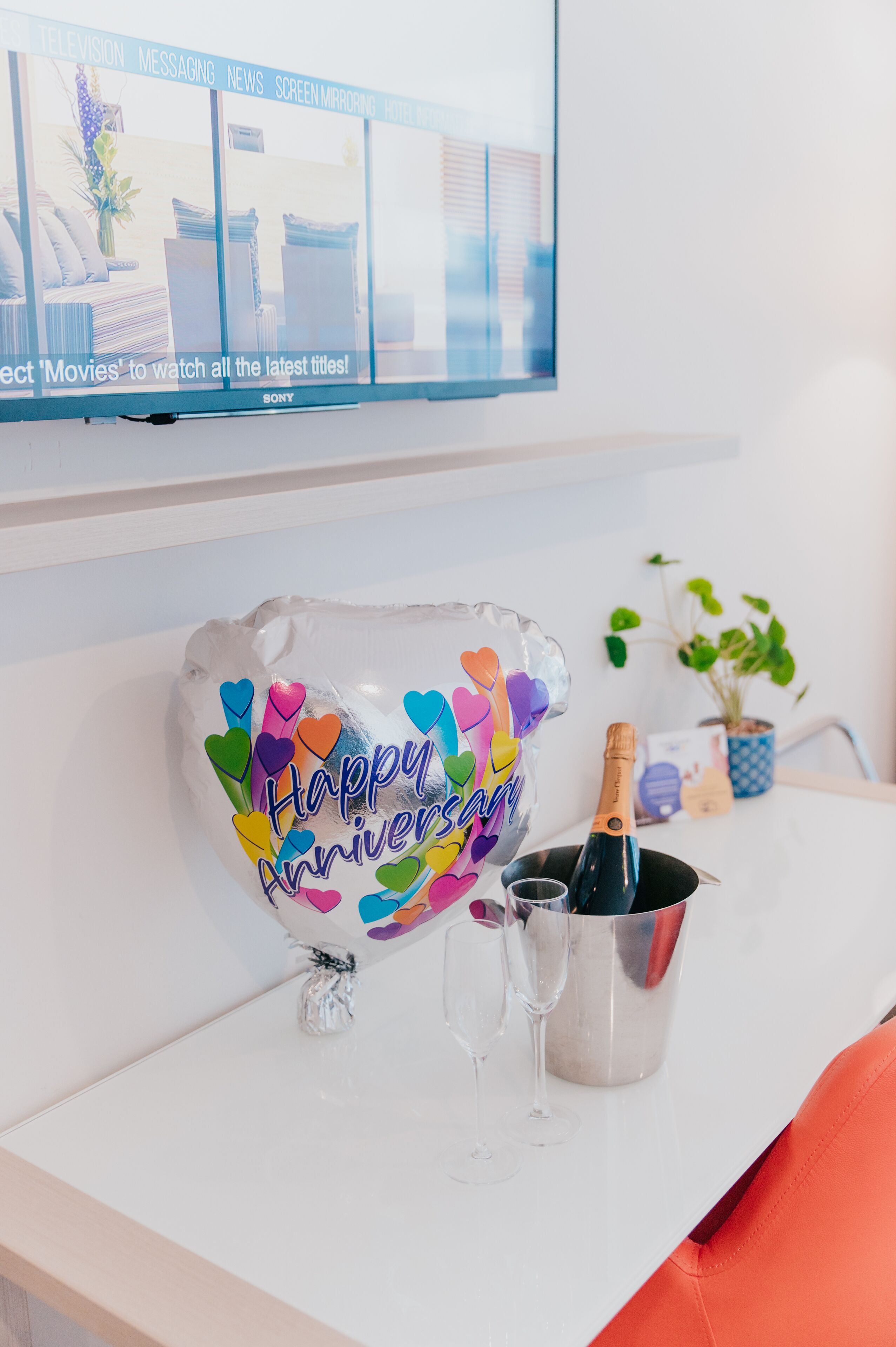 A white table with a colorful balloon, two champagne flutes, and a bottle of champagne.
