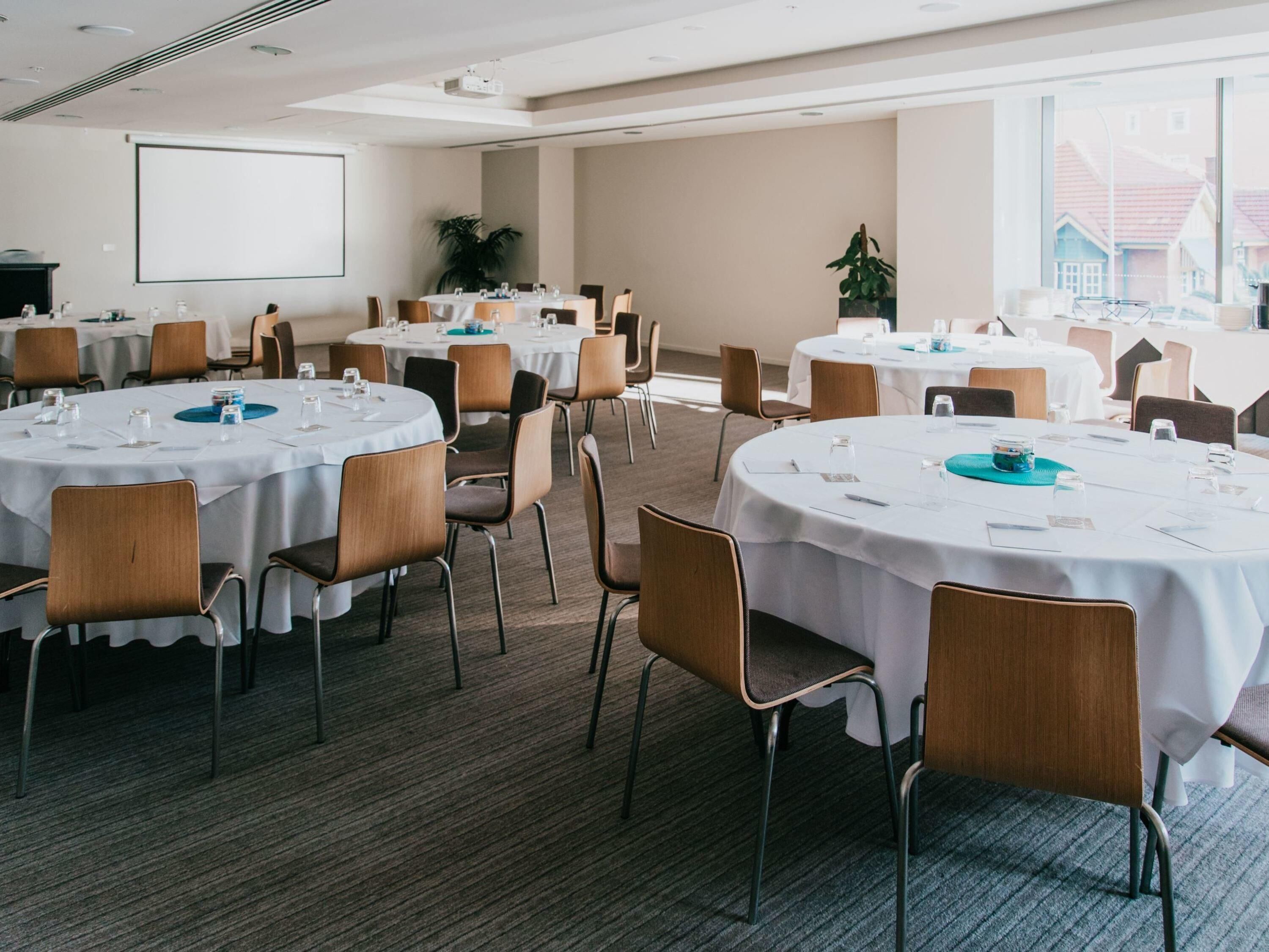 A conference room with multiple tables, chairs, a projector screen, and a large window.