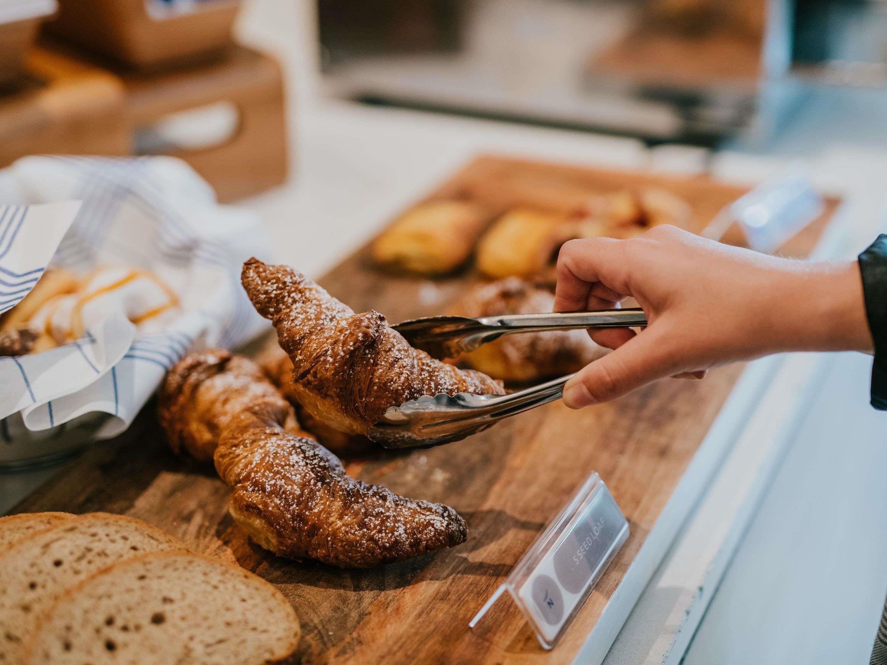 Uma pessoa usando pinças para manusear um doce, com pão e outros produtos de panificação em uma bancada de madeira.