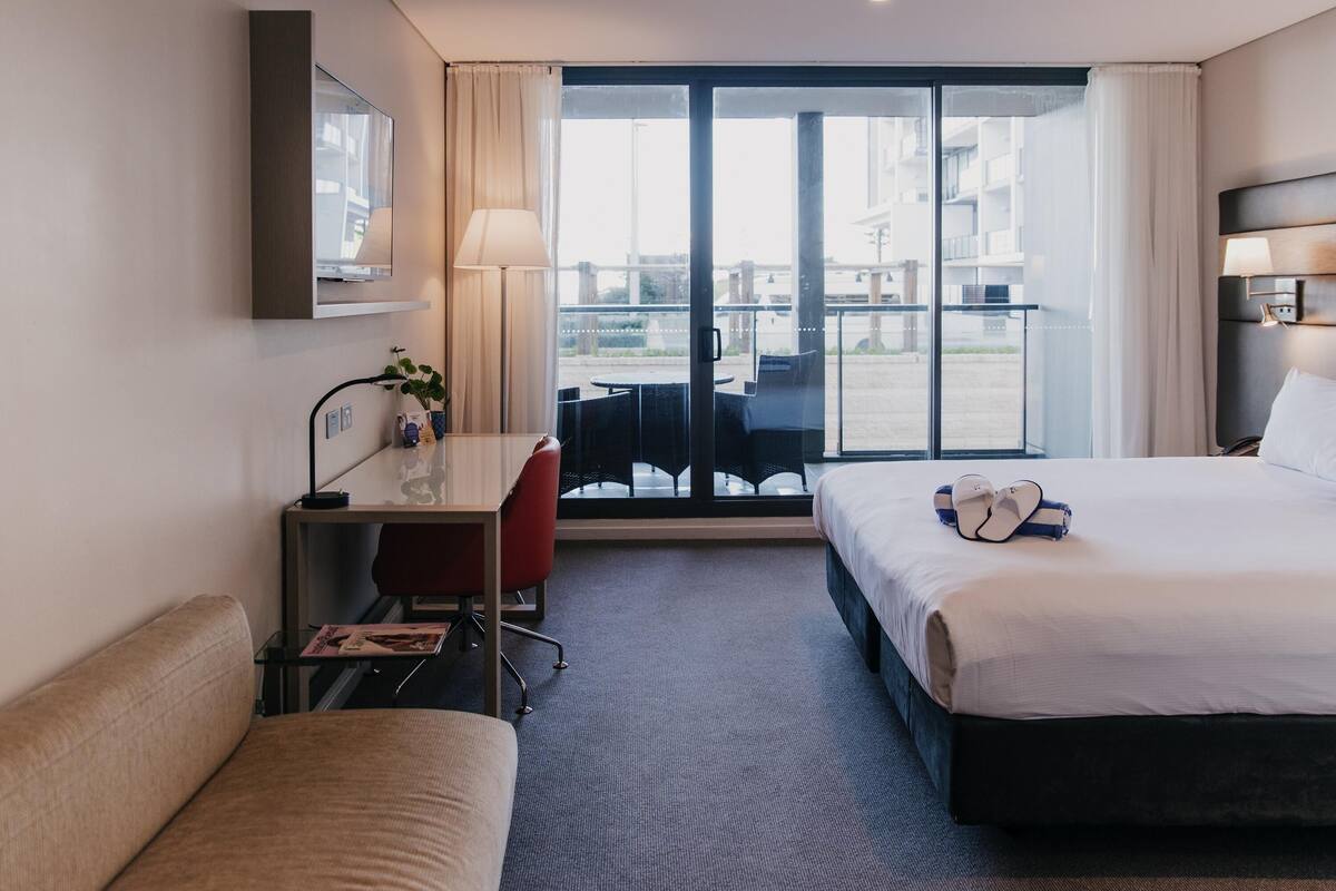 A hotel room with a desk, two beds, a chair, a lamp, and a view of the outside.