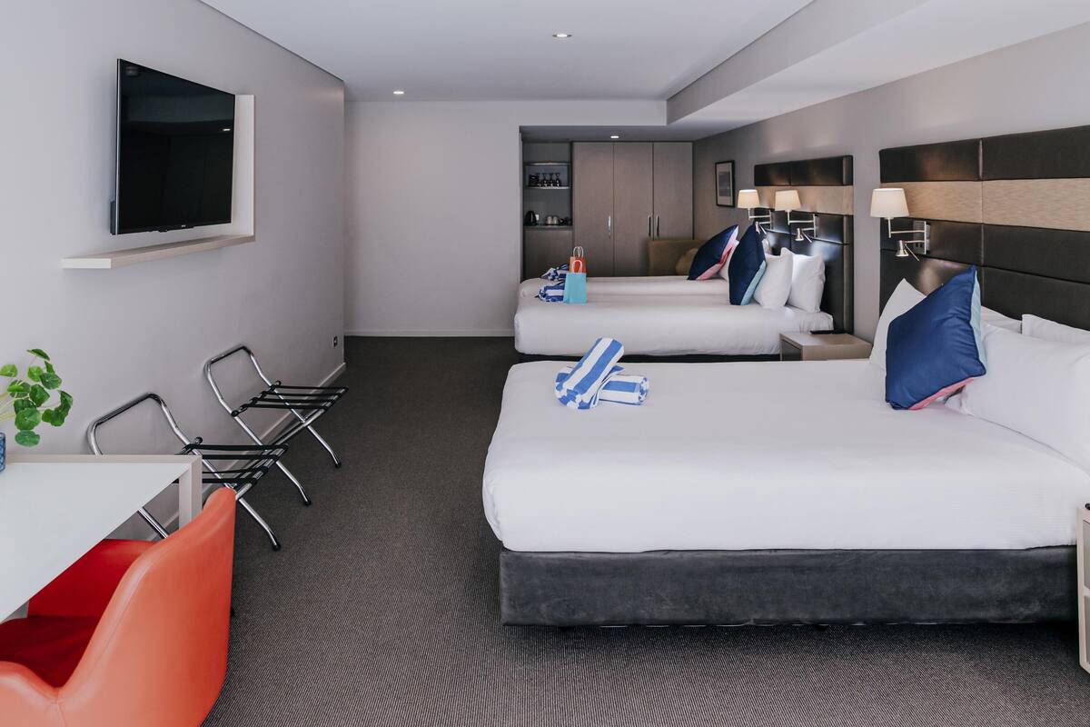 A hotel room with two beds, a desk, chairs, a TV, and a kitchenette.