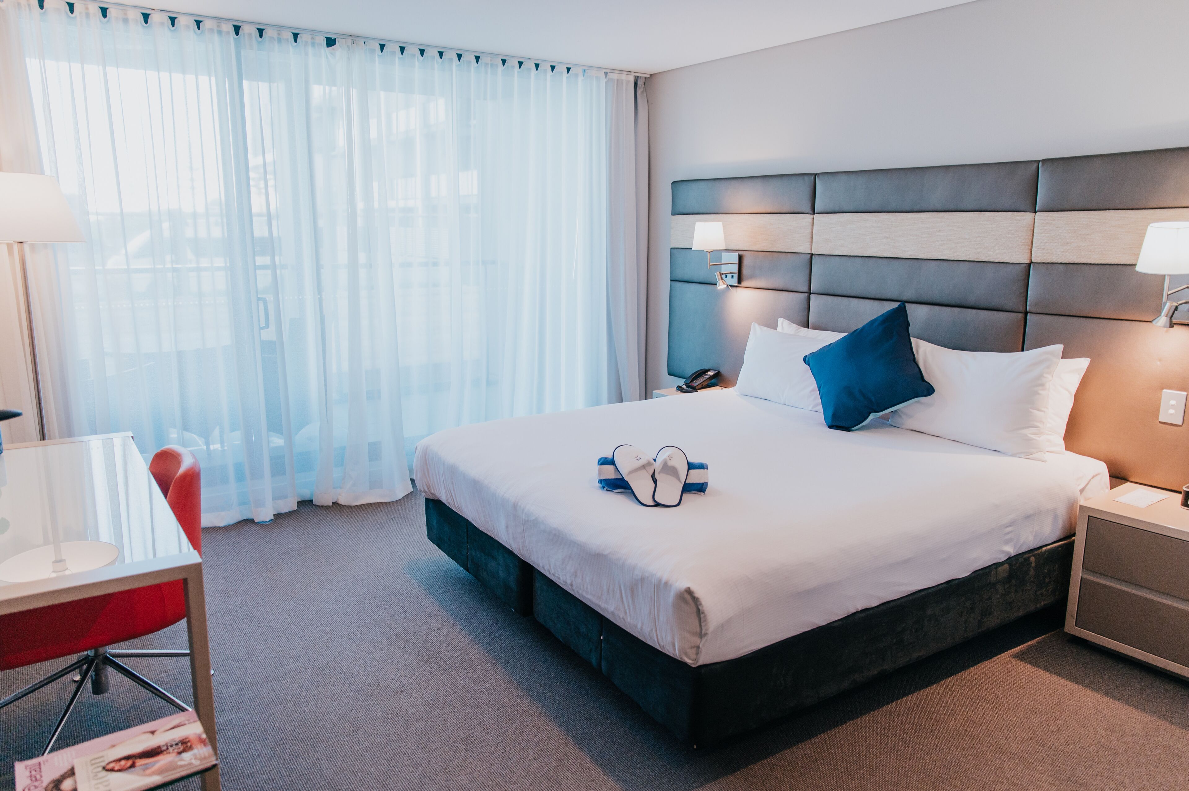 A hotel room with a large bed, a bedside table, a desk with a chair, and a window with sheer curtains.
