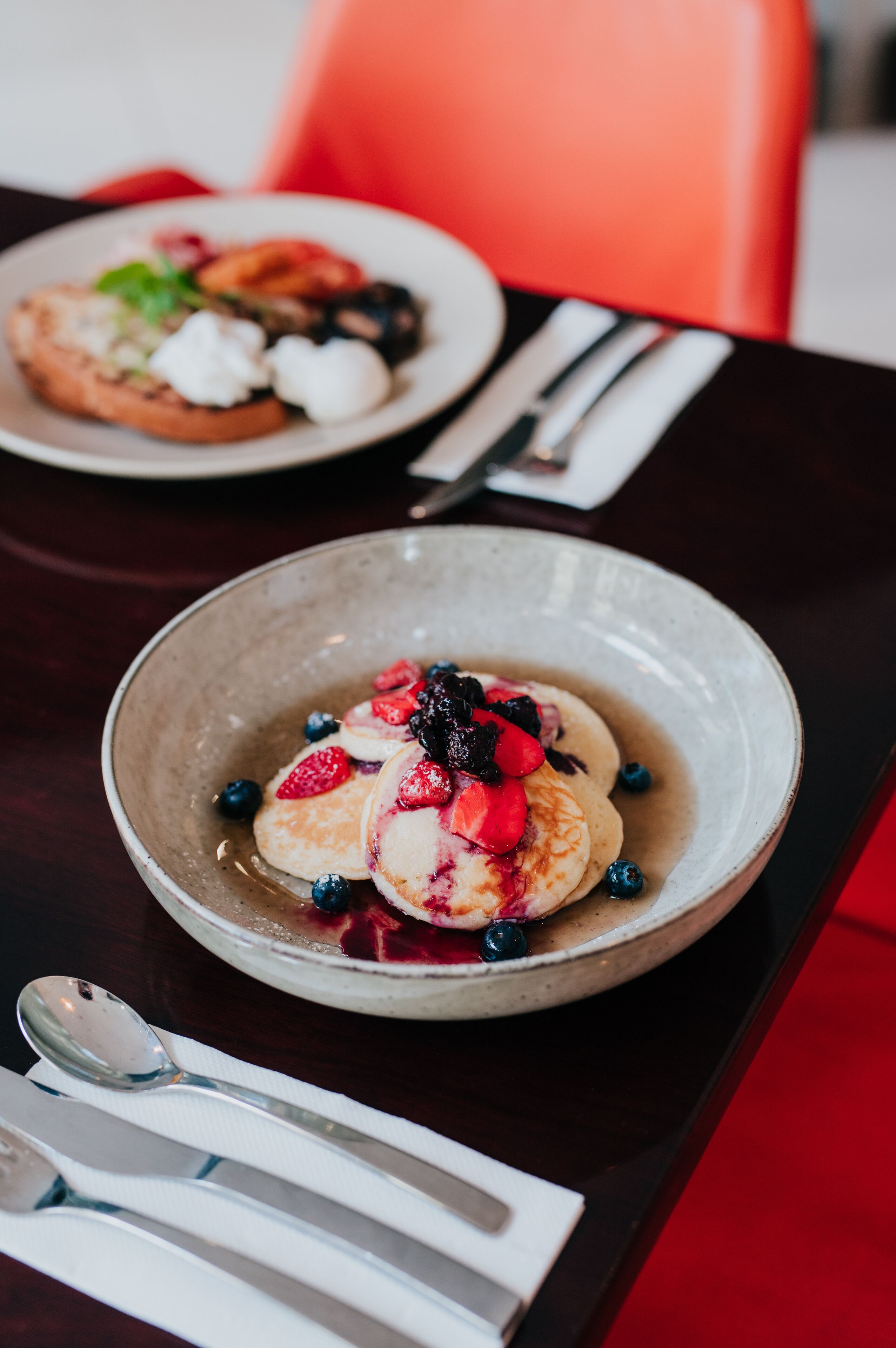 A dining table with a plate of pancakes topped with berries and syrup, a spoon, and a fork on a napkin.