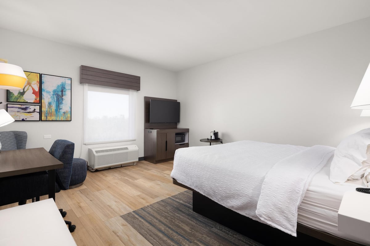 A hotel room with a bed, a desk, a chair, a TV, and artwork on the wall.
