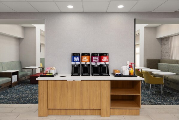 Un espace dédié au café, équipé de quatre distributeurs étiquetés « Café torréfié foncé », « Café torréfié léger », « Café décaféiné » et « Eau chaude ». Des coins salon avec des tables et des chaises sont aménagés autour de cet espace.
