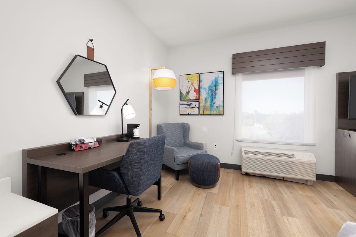 A modern hotel room with a wooden desk, a blue office chair, a grey armchair, a large mirror, a floor lamp, and a window with a view.
