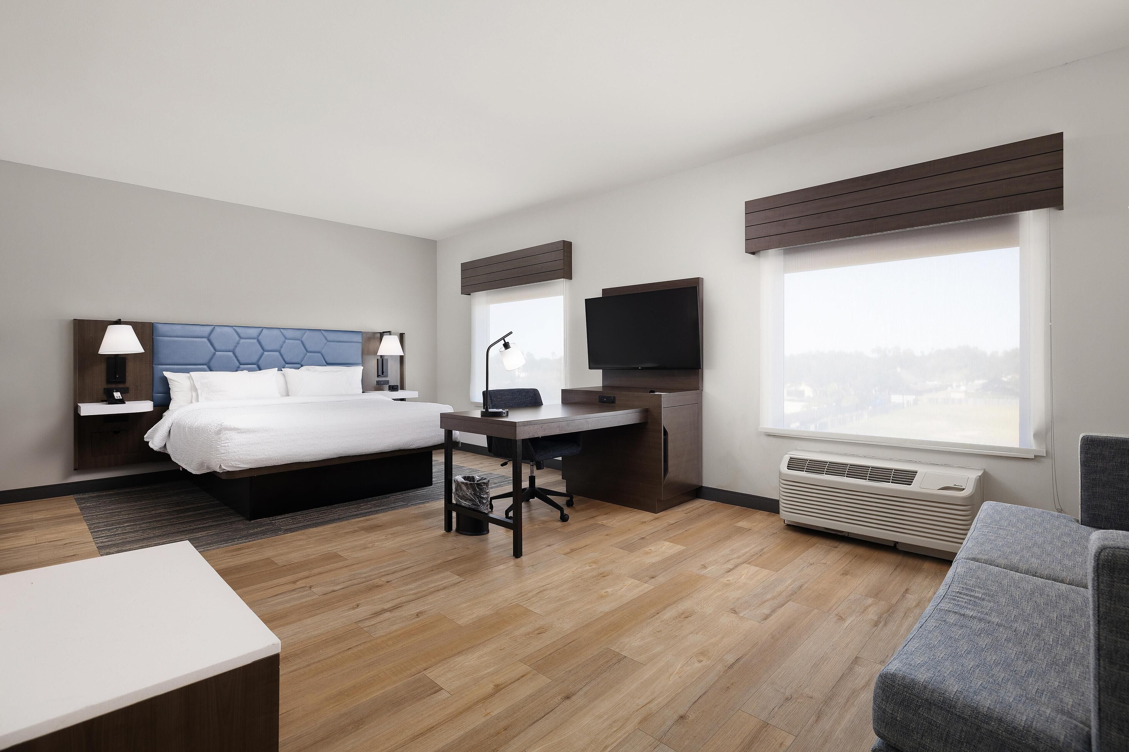 A modern hotel room with a bed, desk, chair, TV, and large windows.