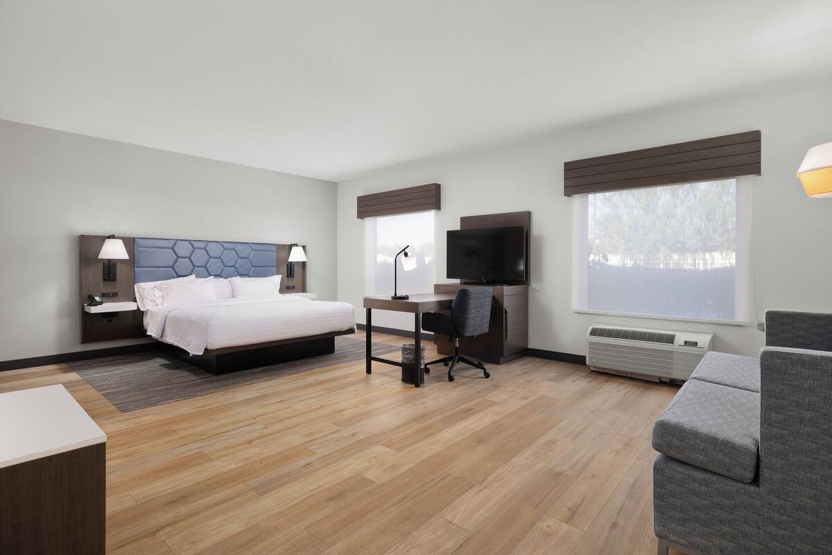 A hotel room with a bed, desk, chair, TV, and large windows.