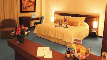 A hotel room with a large bed, a desk with a computer, a chair, a small table with a vase of flowers, and a minibar.