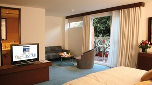 A hotel room with a bed, a sofa, a chair, a TV, and a balcony with plants. - Blue Suites Hotel (Bogotá)