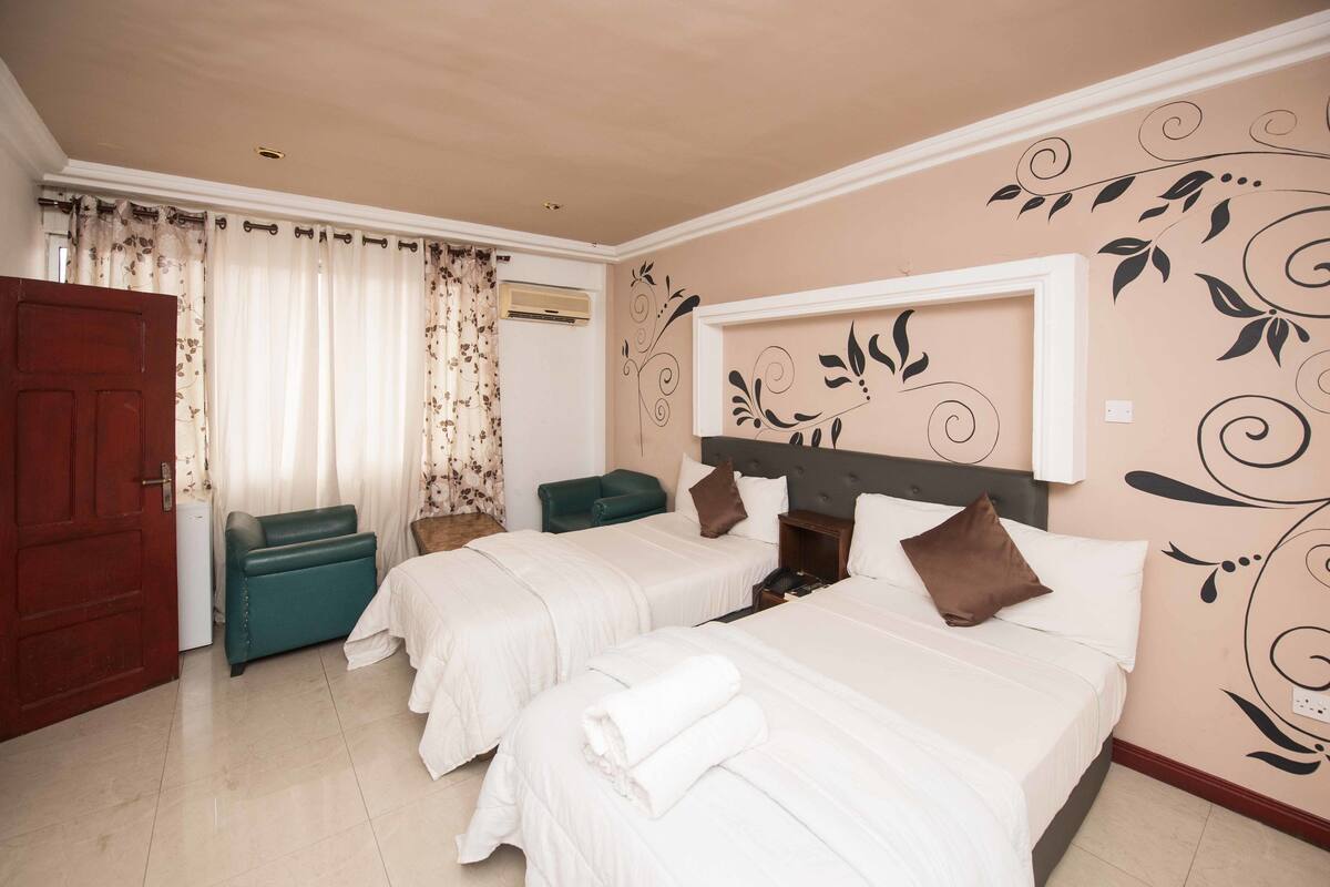 Habitación de hotel con dos camas, un sofá verde, una mesita de noche de madera y una pared decorativa con motivos florales.