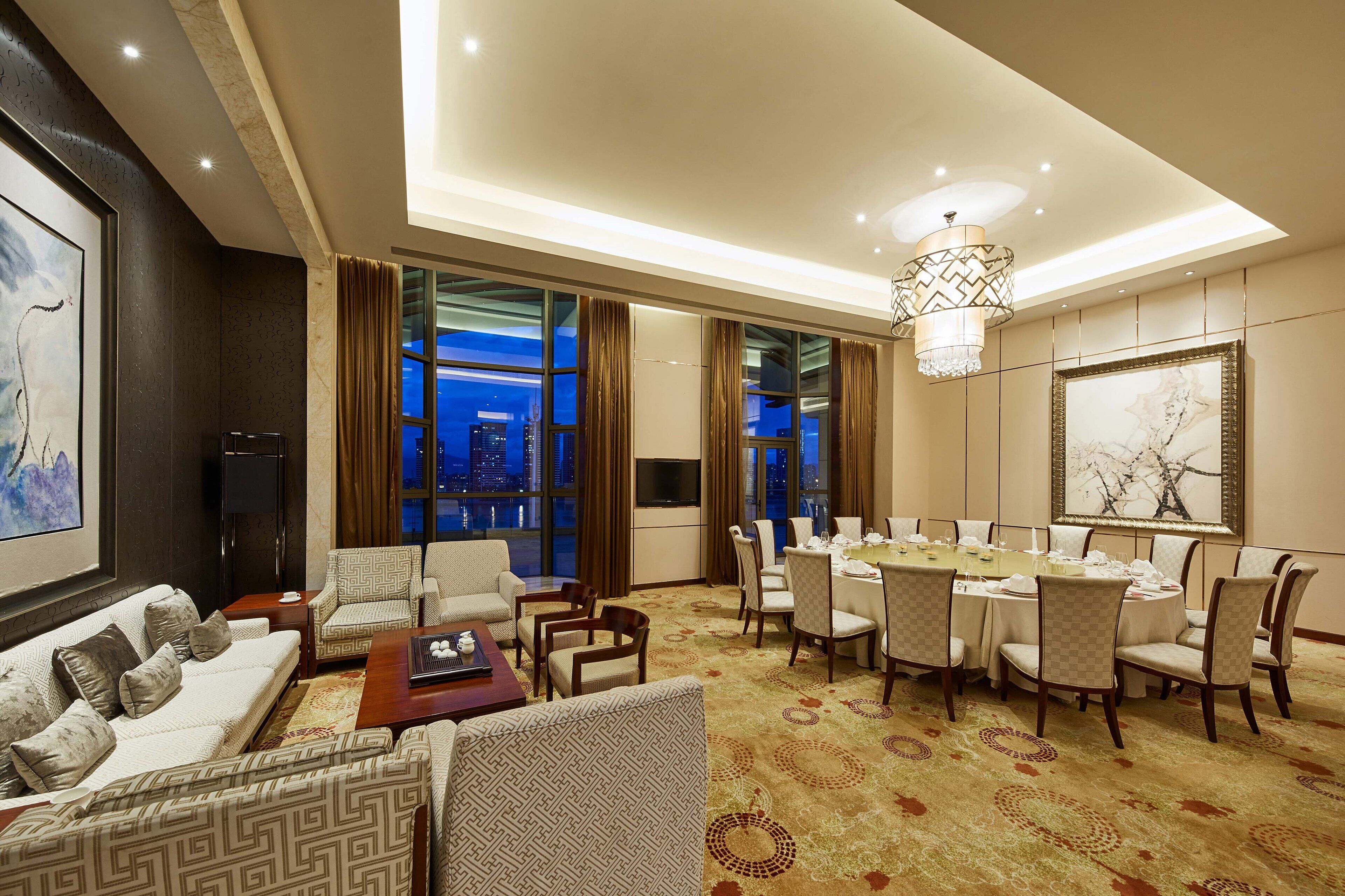 A hotel room with a living area, dining setup, and large windows with a city view.