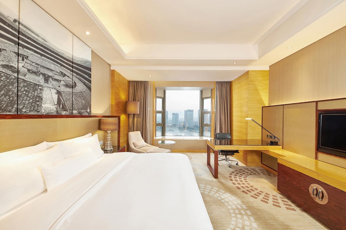 Ein Hotelzimmer mit einem großen Bett, einem Schreibtisch, einem Sessel, einem Fernseher und Blick auf die Stadt.