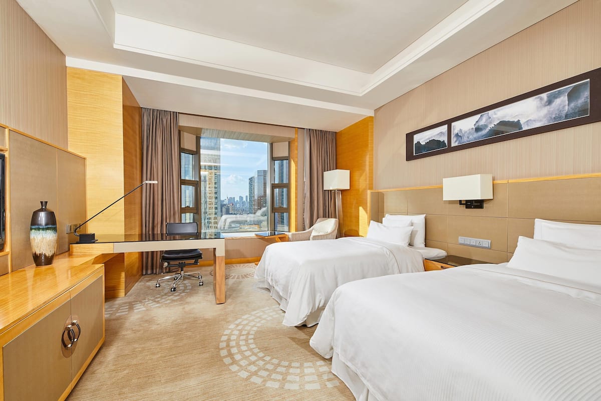 Ein Hotelzimmer mit zwei Betten, einem Schreibtisch, einem Stuhl, einer Lampe und einem großen Fenster mit Blick auf die Stadt.