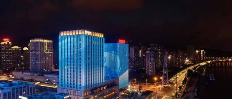 A high-rise hotel with a prominent blue-lit facade, situated by a waterfront with a busy road and other buildings in the background.