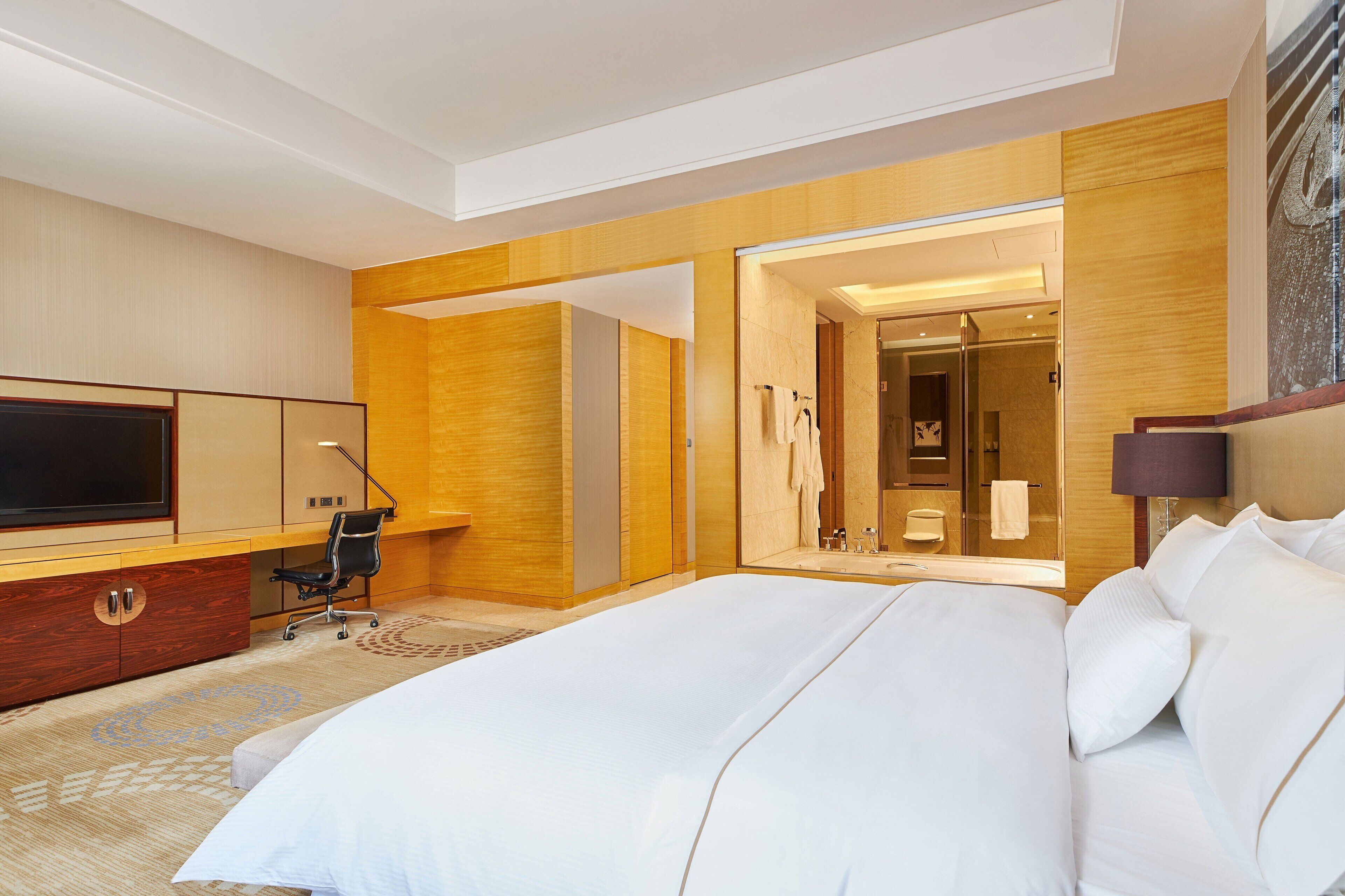 A modern hotel room with a large bed, a desk with a flat-screen TV, a chair, and a bathroom visible through an open door.