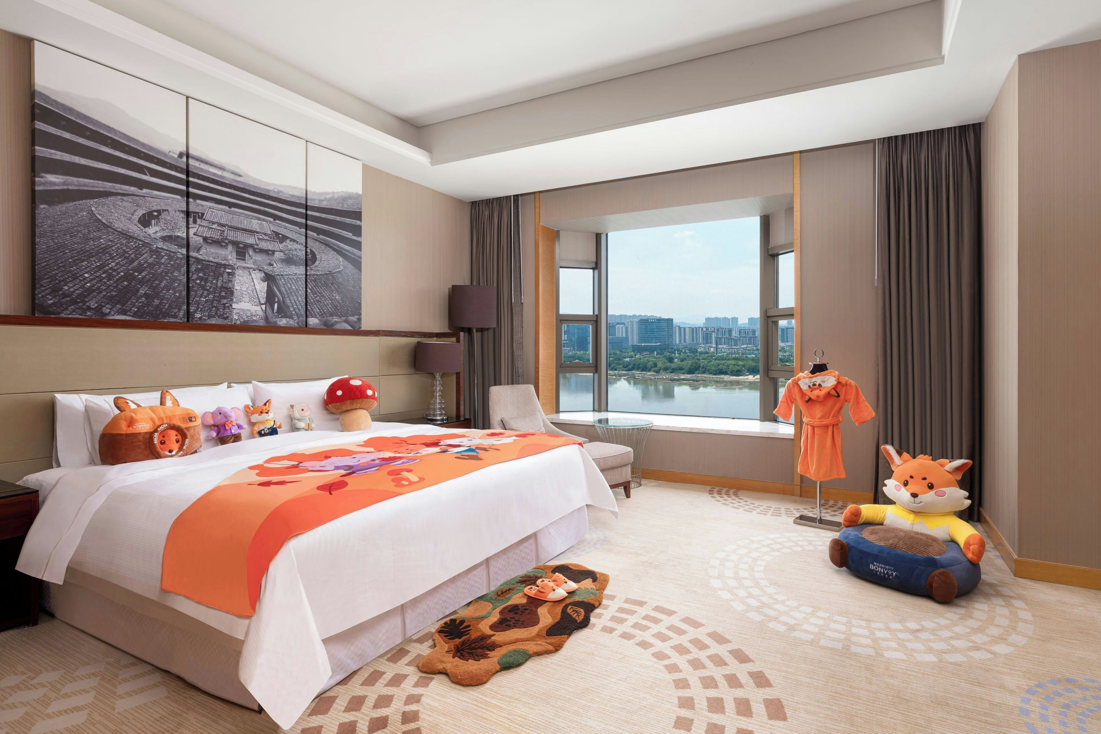 A hotel room with a large bed, a city view, a painting on the wall, and various plush toys.