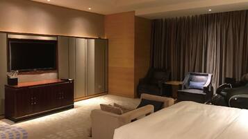 A hotel room with a large bed, a TV on a wooden stand, a seating area with a chair and a small table, and a large window with curtains.