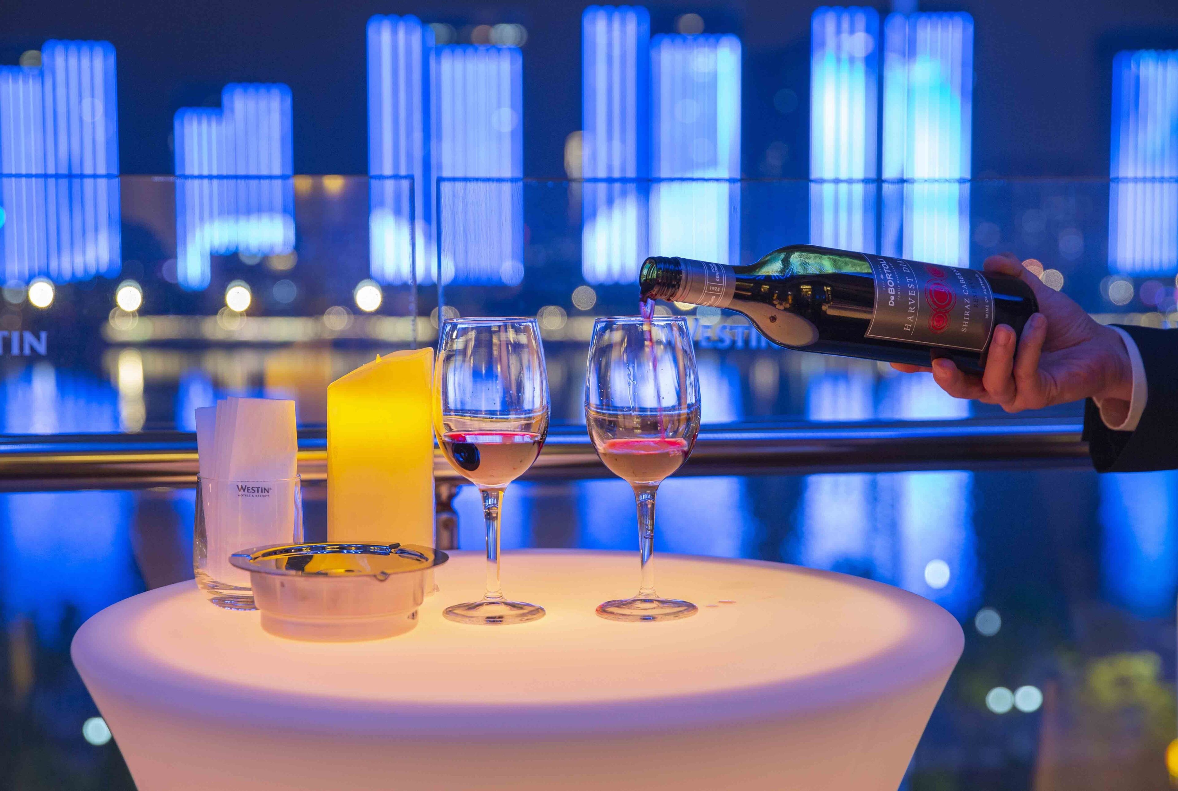 A hand pouring wine into glasses on a table with a cityscape view in the background.