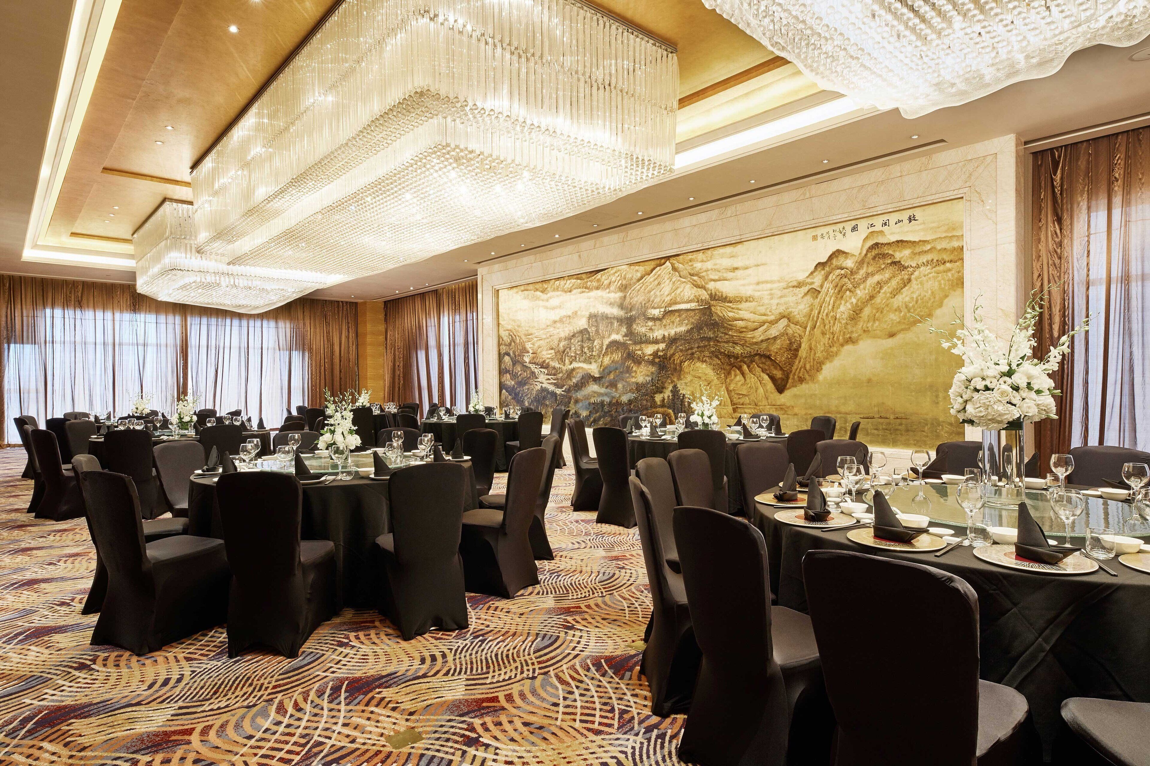 A banquet hall with round tables set for dining, a large mural on the wall, and a chandelier.
