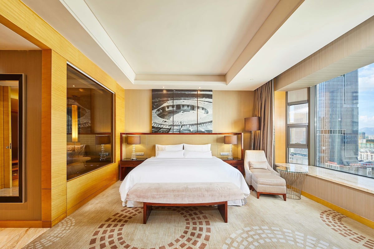 Ein modernes Hotelzimmer mit einem großen Bett, einer Sitzecke, einem großen Fenster mit Blick auf die Stadt und einem großen Kunstwerk an der Wand.