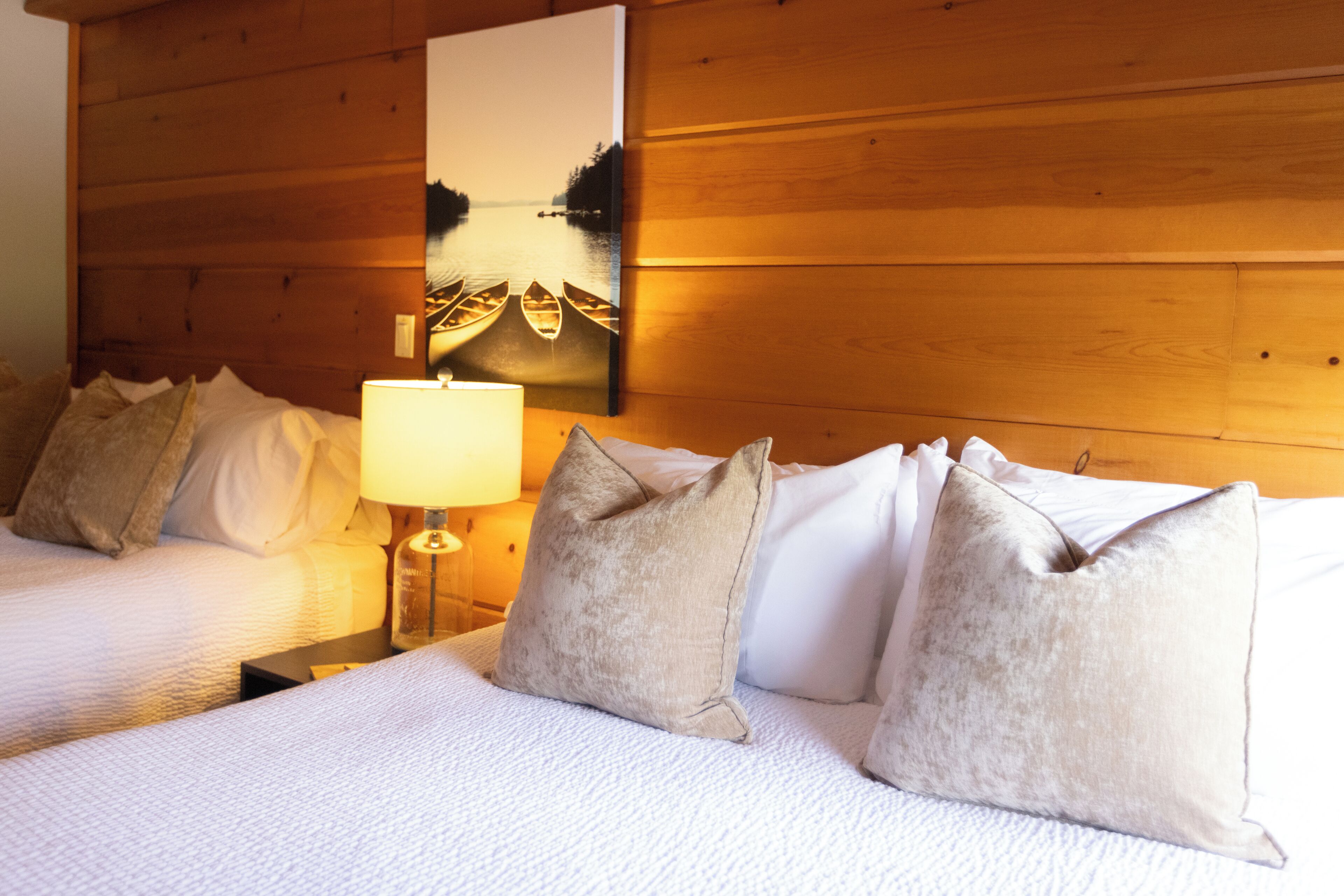 A hotel room with two beds, a wooden headboard, a nightstand with a lamp, and a painting of boats on water.