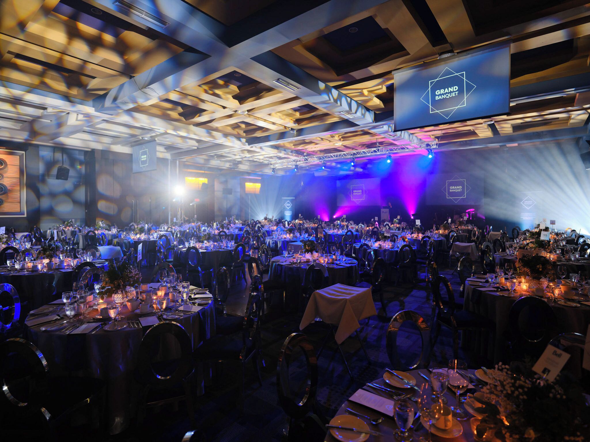 A banquet hall with tables set for a formal event, featuring a high ceiling with geometric patterns and a stage with lighting.